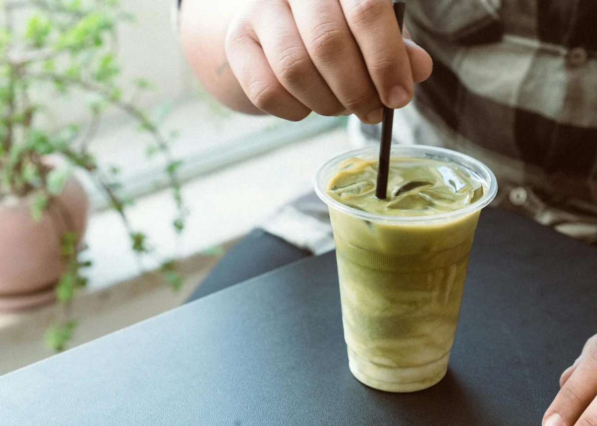 Ruka meša zeleni smoothie slamčicom u providnoj plastičnoj čaši, biljka u pozadini