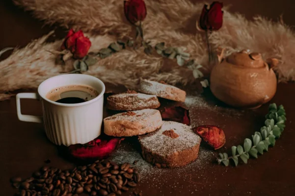 Bela šolja crne kafe, okrugli kolačići u prahu i sušene ruže na tamnoj rustičnoj podlozi