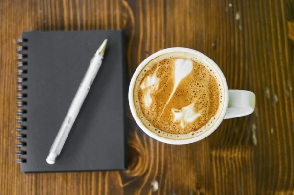 Bela šolja kafe s latte art uzorkom pored crne beležnice i olovke na drvenoj površini