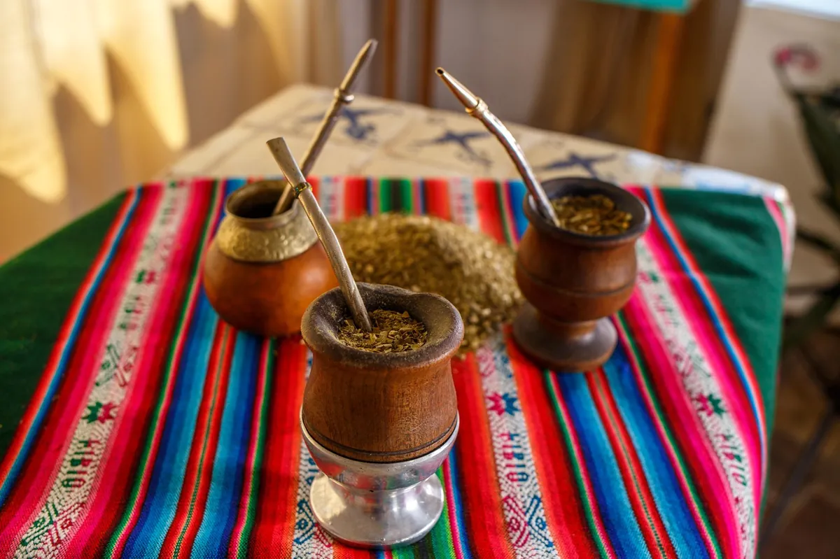 Tri drvene tikvice sa metalnim bombiljama i rasutim sušenim listovima yerba mate na šarenom tradicionalnom tekstilu